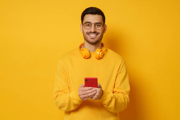 istock Young man in glasses, looking at camera with happy smile, holding phone in hands, wearing headphones round neck, feeling relaxed, isolated on yellow background 1237258343