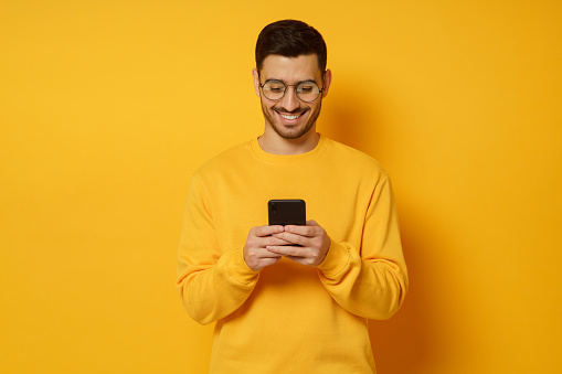 Joven hombre de moda con gafas y suéter, sintiéndose feliz por la conversación en mensajes de texto con amigos por teléfono, sonriendo mientras mira la pantalla, aislado en el fondo amarillo photo