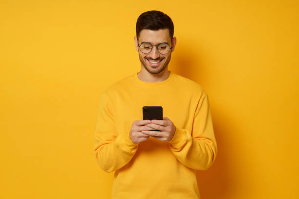 istock Young trendy man in glasses and sweater, feeling happy about conversation in text messages with friends via phone, smiling while looking at screen, isolated on yellow background 1237258345