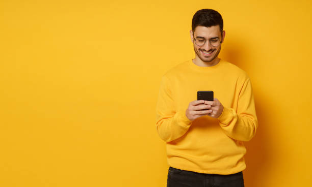 istock Horizontal banner of guy exchanging text messages with friends, smiling happily and feeling relaxed, wearing bright sweatshirt, isolated on yellow background, copy space on left 1237258350