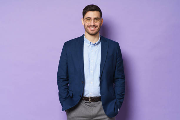 istock Business guy dressed in jacket and shirt, wearing glasses, standing in confident relaxed pose, hands in pockets, smiling happily, isolated on purple background 1237258363