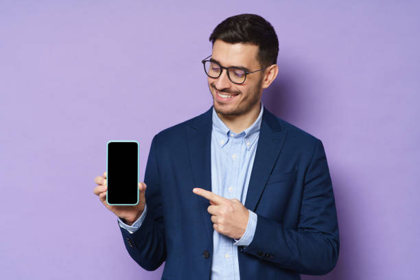 ジャケット、シャツ、眼鏡の若いビジネスマンは、空白の画面でスマートフォンを保持し、コピースペースが含まれ、それを指し示し、紫色の背景に隔離された高品質の金融アプリをアドバ� - 見せる ストックフォトと画像