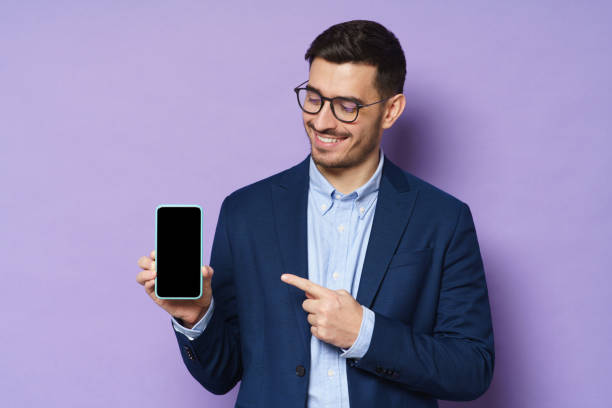 jovem empresário de jaqueta, camisa e óculos segurando smartphone com tela em branco, espaço de cópia incluído, apontando para ele, aconselhando aplicativo de finanças de alta qualidade, isolado em fundo roxo - men blank holding showing - fotografias e filmes do acervo