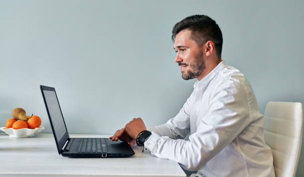 istock Side view indoor image of smiling bearded man working on laptop for conversation online with friends, browsing online. Handsome male sitting at the desk in the kitchen, working on website on computer. 1237314304