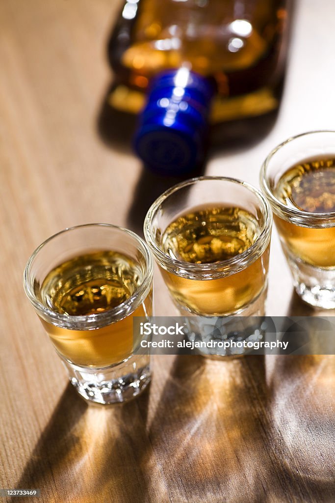 Shots of whiskey three glasses full of dark colored alcohol on top of a bar table along with a bottle of whiskey. The shot could be whiskey, dark rum, tequila, cognac, or bourbon. Alcohol - Drink Stock Photo