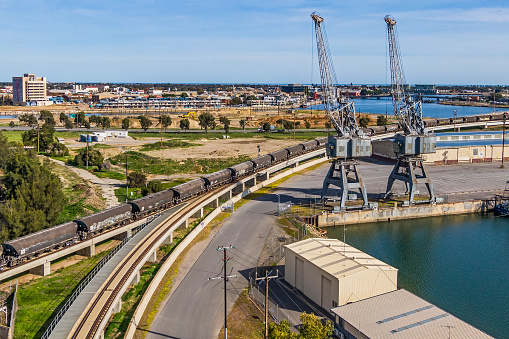 Rail freight grain cars (wagons, hoppers) crossing Port River bridge, past two historic gantry cranes contrasting with new Port Adelaide waterfront housing development in background.
