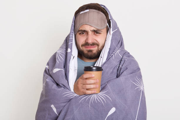 istock Bearded man with curved face holding coffee or tea, can not wake up in morning, guy wrapped blanket, unshaven man against white wall, being in bad mood. 1237453223