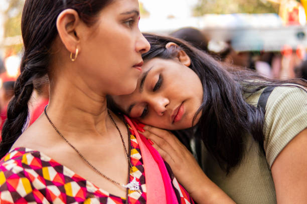 Teenager daughter resting her head on mothers shoulder and thinking with blank expression while standing in market at day time.