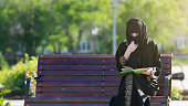 Muslim woman is reading a book on a bench.