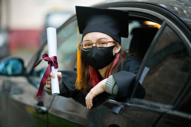 teenage girl wearing graduation gown and cap and protective mask with diploma in car - university graduation car student imagens e fotografias de stock