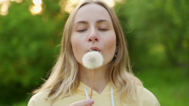 A beautiful young woman stands in the forest and blows a dandelion. On the street. enjoy nature. A healthy, smiling girl on a summer lawn. Allergy-free concept