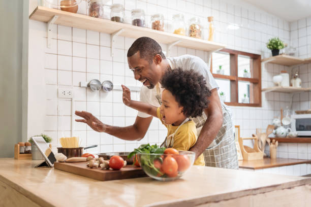 아프리카계 미국인 아버지와 작은 아들이 디지털 태블릿으로 화상 통화를하고 부엌에서 요리하는 동안 가족과 이야기하는 것을 웃으며 손을 흔들었습니다. 흑인 가족은 집에서 전염병 바이러� - 소년 이미지 뉴스 사진 이미지