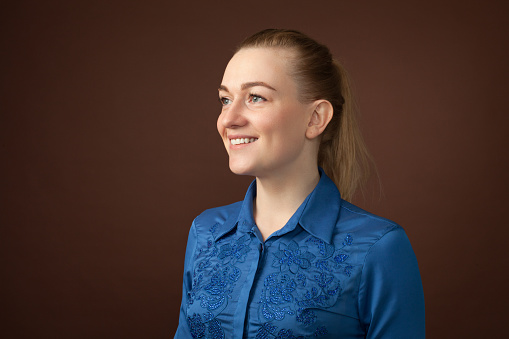 close-up studio portrait of a 30 year old blonde woman with ponytail hair in a blue blouse on a brown background