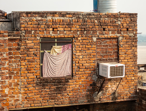 Varanasi, Uttar Pradesh, India - February 2015: The old, bare, brown brick walls of a house with a mesh window and an air conditioner.