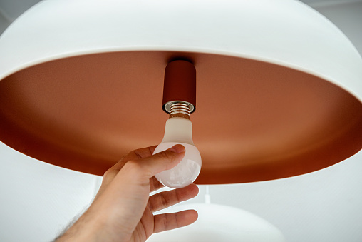 Replacing the light bulb in the luminaire, a close-up of a human hand spins a low-energy led lamp in the shade of frosted glass.