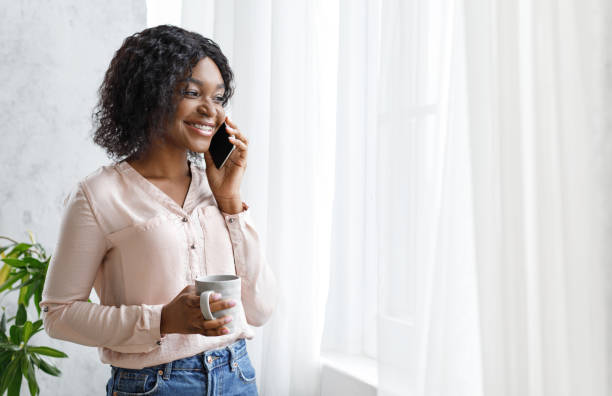 istock Attractive Black Woman Drinking Coffee And Talking On Cellphone Near Window 1237954946