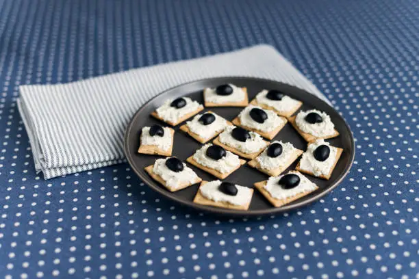 Photo of Plate with crackers with soft cheese and olives on a blue polka dot tablecloth.