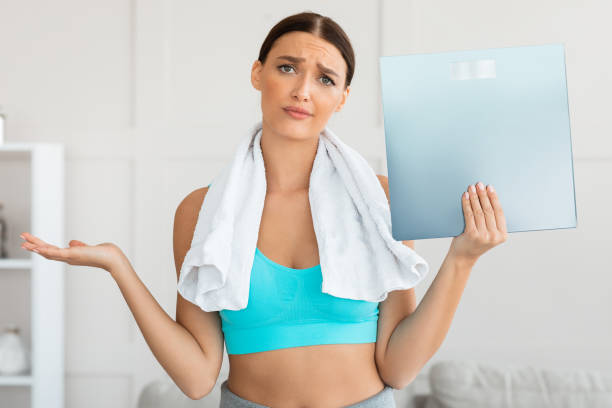 Discontented Girl Holding Weight Scales Standing At Home Discontented Girl Holding Weight Scales Not Losing Kilograms On Unsuccessful Diet Standing At Home. Weight-Loss Failure gaining weight stock pictures, royalty-free photos & images