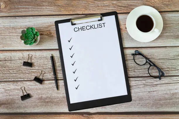 Checklist on blank page with pen, green plant, cup of coffee, eyeglasses and paper clips top view on wooden office or home table