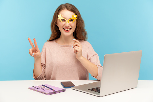 Funny optimistic woman employee covering eyes with bright paper glasses, showing victory gesture and wearing humorous masquerade accessory, having fun at workplace while working in office. indoor