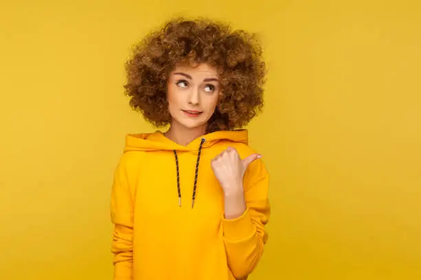 Photo of Look, advertise here! Portrait of pensive perplexed curly-haired woman in urban style hoodie pointing side