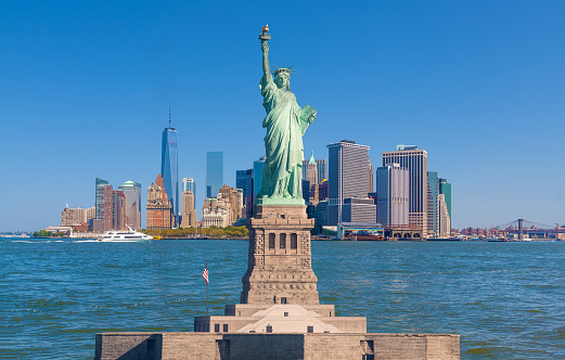 Statue of Liberty and New York City Skyline with Manhattan Financial District, Battery Park, Water of New York Harbor, World Trade Center, Tourboat and Blue Sky.