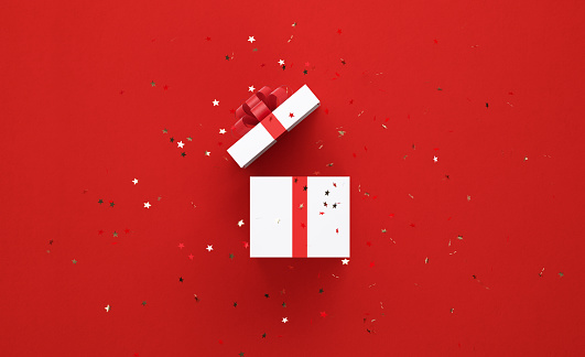 Star shaped confetti falling over white gift box tied with red ribbon on red background. Horizontal composition with copy space, Great use for Christmas and Valentine's Day related gift concepts.