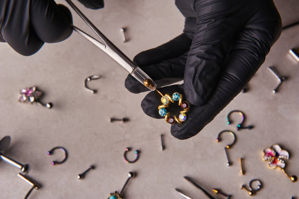 piercer master hand in einem medizinischen schwarzen handschuh hält einen ohrring. ohrringe liegen herum - piercing stock-fotos und bilder