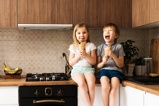 Two little kids eating ice cream in kitchen at home, smiling and enjoying, lifestyle