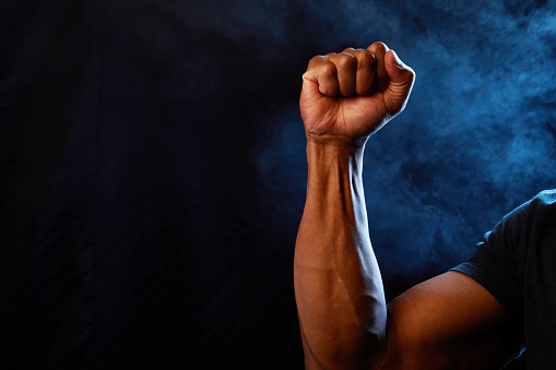 Studio image of an African American fist held up in strength, unity, and defiance with smoke background and blue lights representing police.