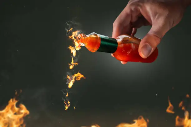 A small bottle with burning hot chili sauce pouring out of it  held by a hand. The drops of sauce are actually on fire.