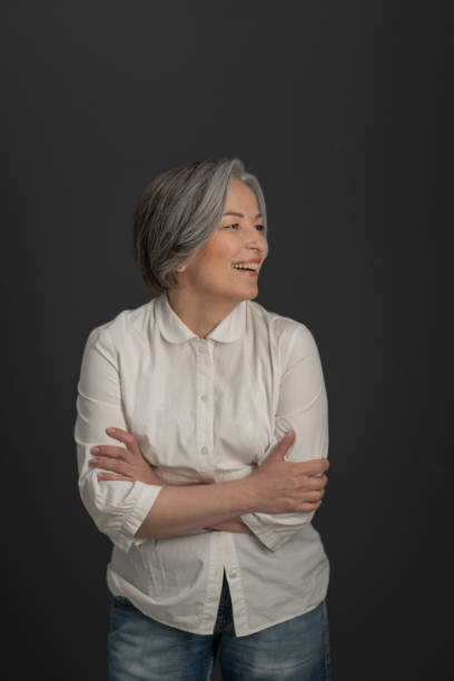 laughing charming woman looks away confusedly with her arms folded. joyful mature woman posing in studio on gray studio background - senior getting groomed studio imagens e fotografias de stock