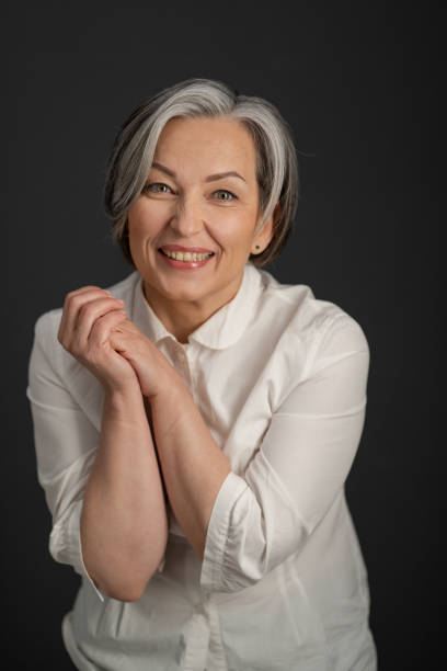 istock Joyful Caucasian woman smiles broadly while looking at camera and clasped her hands. Enthusiastic gray-haired mature woman in a white shirt posing in studio 1238350699