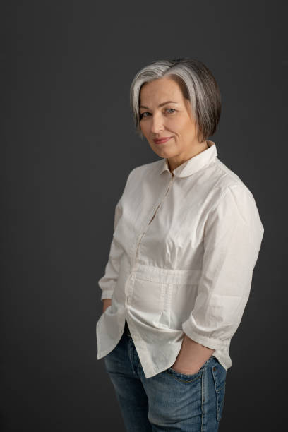 istock Gray-haired mature woman smiles holding hands in pockets while looking at camera. Charming caucasian woman in white shirt posing on a gray background 1238371726