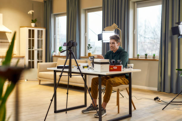 istock Bringing Joy. Young cheerful male technology blogger smiling, while recording video blog or vlog about new camera lens and other gadgets sitting at home studio. Blogging, Work from Home concept 1238414799