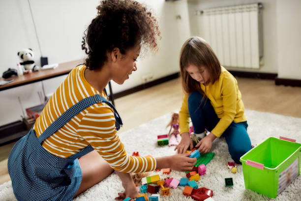 viel spass. kaukasische süße kleine mädchen verbringen zeit mit afrikanischen amerikanischen baby-sitter. sie spielen mit bauspielzeug-set, sitzen auf dem boden - babysitter stock-fotos und bilder