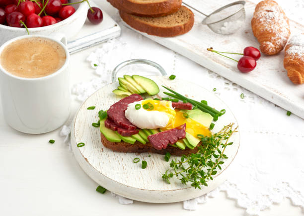 toast with poached egg and avocado on a round board, next to croissants and ripe red cherries, morning breakfast, top view on a white table