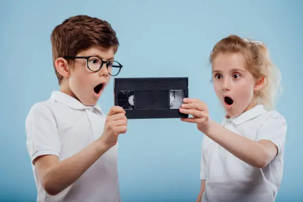 surprised little boy and girl with video tape, isolated on blue background, old gadgets