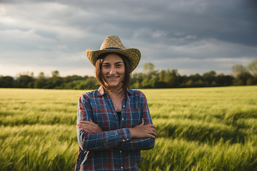 Portrait of a young and beautiful woman farmer with farmer hat standing with cross arms in the middle of the wheat field at sunset. She is smiling.