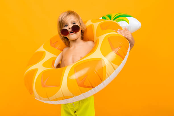 portret zabawnego europejskiego blond chłopca w żółtych kąpielówkach i okularach przeciwsłonecznych z ananasem na kręgu pływackim na pomarańczowej ścianie - swimming tube inflatable circle zdjęcia i obrazy z banku zdjęć