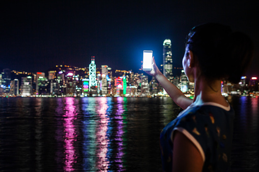 Young Chinese woman holding mobile phone at Victoria Harbour in Hong Kong at night