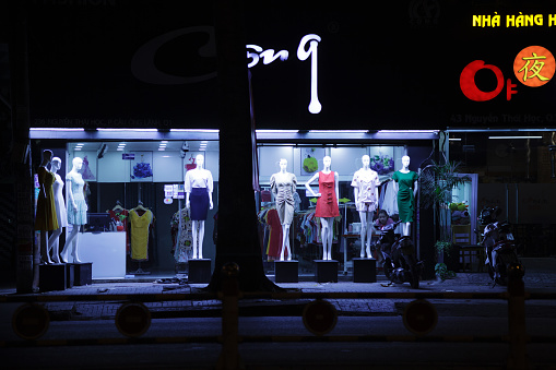 Ho Chi Minh City, Vietnam - May 8, 2020: Women's dresses displayed on dummies in front of a fashion store. Young Asian saleslady alone in a clothing boutique at night