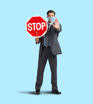 A man wearing a protective mask holds out his hand while he holds a stop sign in front of him against a blue background.