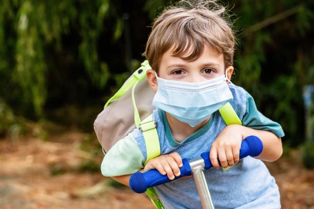 little boy riding a push scooter carrying a back pack wearing a protective face mask - little boys preschooler back to school backpack imagens e fotografias de stock