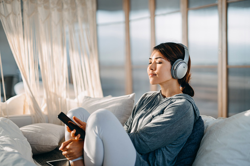 Joven asiática relajada con los ojos cerrados sentados en su cama disfrutando de la música sobre los auriculares desde el teléfono inteligente en casa photo