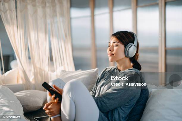 Relaxed Young Asian Woman With Eyes Closed Sitting On Her Bed Enjoying Music Over Headphones From Smartphone At Home Stock Photo - Download Image Now