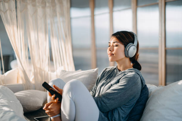 relaxed young asian woman with eyes closed sitting on her bed enjoying music over headphones from smartphone at home - ouvir musica imagens e fotografias de stock