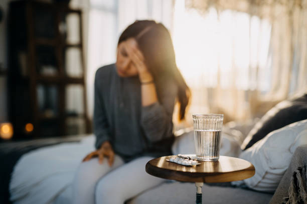 joven asiática sentada en la cama sintiéndose enferma y sufriendo de dolor de cabeza, un vaso de agua y medicinas en la mesa auxiliar - pill human pregnancy capsule women fotografías e imágenes de stock