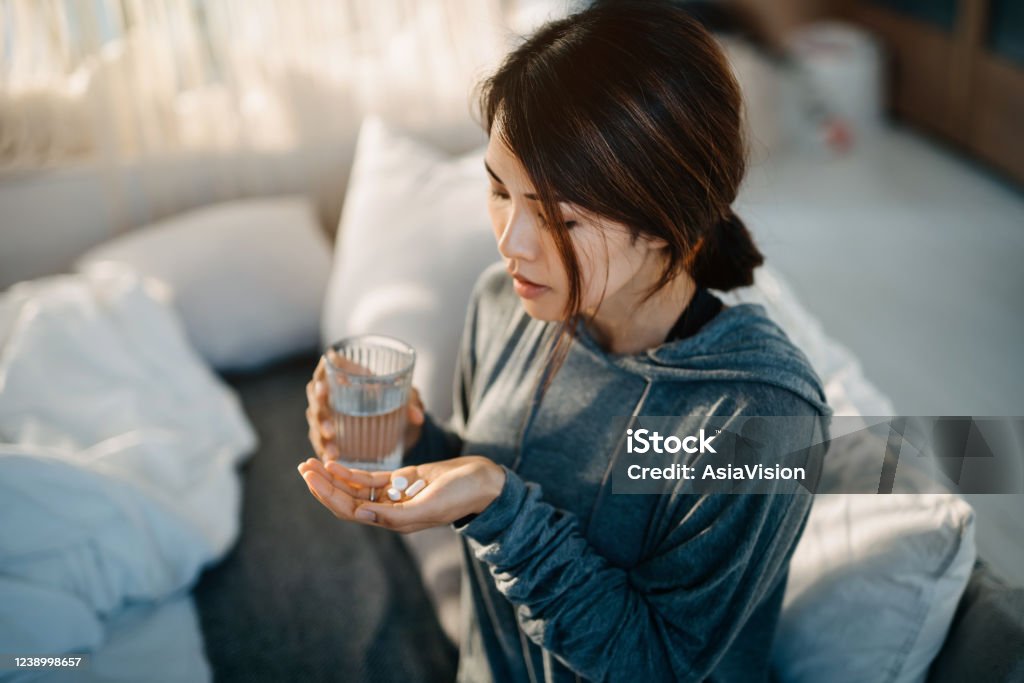 Young Asian woman sitting on bed and feeling sick, taking medicines in hand with a glass of water Headache Stock Photo