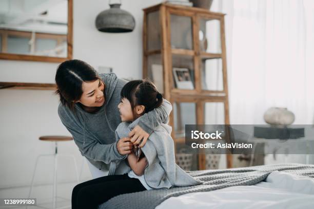Caring Young Asian Mother Putting A Coat On Her Daughter At Home Stock Photo - Download Image Now